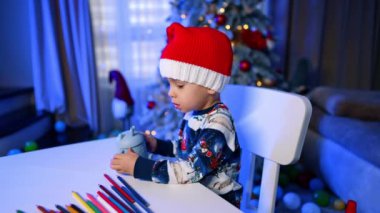 Noel Baba şapkalı sevimli beyaz çocuk pipetli komik bir bardaktan içiyor. Çocuk içkisini bitirirken bardağı sallıyor. Arkaplanda bulanık bir Noel ağacı.