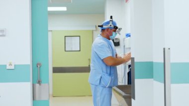 Mavi üniformalı doktor, şapka, maske ve ameliyat öncesi odasında duruyor. Cerrah ellerini kurutmak için peçete kullanıyor ve dezenfektan sürüyor..