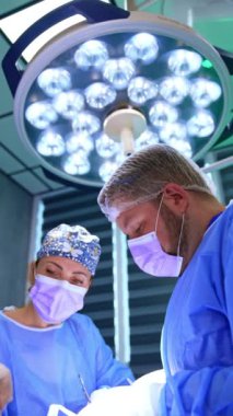 Kafkas cerrahlar, modern ameliyathanede yuvarlak lambayla aydınlatılmış bir operasyonda çalışıyorlar. Doktorlar tiroid üzerinde işlem yaparken elektrikli cihaz ve metal aletler kullanırlar. Düşük açı. Dikey video.