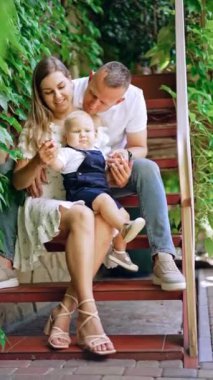 Anne ve baba sevgili bebekleriyle merdivenlerde oturuyorlar. Küçük çocuk ayağa kalkmaya çalışırken hareket etmeye başlıyor. Dikey video.