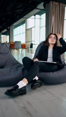 Geniş bir ofisteki fasulye çuvalında oturan genç bir kadın. Bayan kucağında bir dizüstü bilgisayar tutuyor. Uzaktan çalışma konsepti. Düşük açı. Dikey video.