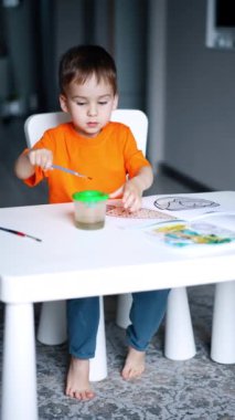 Evde turuncu tişörtlü yaratıcı çocuk resim yapıyor. Küçük çocuk hobisine odaklanmış. Dikey video.