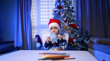 Kırmızı Noel Baba şapkalı çocuk komik bir bardaktan içiyor. Bebek içtikten sonra bir fincan sallar. Arkaplanda Noel ağacı.