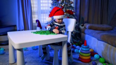 Noel Baba şapkalı sevimli küçük çocuk parlak boncuklarla oynuyor. Çocuk bir Noel süsü düşürür ve almak için ayağa kalkar..