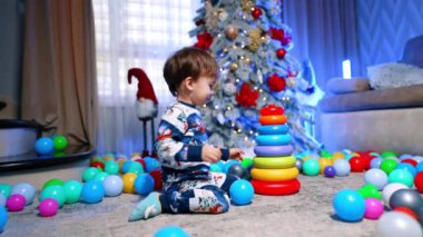 Güzel Noel ağacının yanında oturan neşeli bebek. Gülümseyen çocuk bir piramit yapar ve onu yere atar..