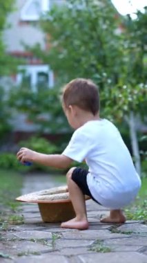 Yalınayak küçük çocuk bahçedeki kaldırım yolunda yatan hasır bir şapkaya doğru koşar. Elinde küçük bir top olan çocuk şapkasını takıp kaçtı. Dikey video.