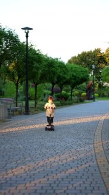 Küçük çocuk mobiletiyle kameraya yaklaşıyor. Sevimli gülümseyen mutlu çocuk gün batımında parkta eğleniyor. Dikey video.