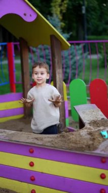 Sevimli çocuk kirli ellerini kum havuzunda gösteriyor. Küçük çocuk kameraya gelip kuma bakıyor. Dikey video.