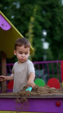 Koyu saçlı erkek bebek dışarıda kumla oynuyor. Çocuk bir kürekle kumu hareket ettiriyor. Dikey video.