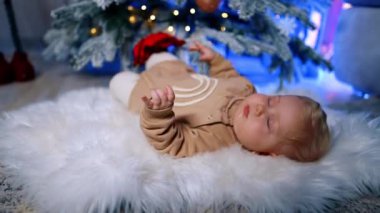 Tatlı sarışın Kafkasyalı bebek Noel ağacının altındaki yumuşak ekose üzerinde yatıyor. Şirin çocuk ağaca dokunuyor..