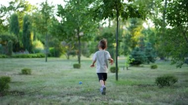 Gri tişörtlü küçük çocuk topunu takip ederek çimlerin üzerinde koşuyor. Küçük çocuk bir oyuncak seçiyor ve kameraya mutlu bir şekilde yaklaşıyor..