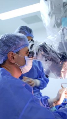Beyin cerrahı, aletleri kullanarak mikroskobun dürbününe aynı anda bakar. Ameliyat odasında çalışan profesyonel bir doktorun yan görünüş portresi. Dikey video.