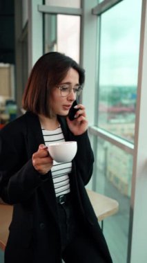 Neşeli Kafkasyalı esmer kahve içiyor ve telefonda konuşuyor. Bayan masaya oturup pencereye bakıyor ve akıllı telefondan konuşuyor. Dikey video.