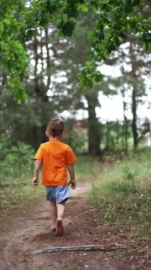 Turuncu tişörtlü çıplak ayaklı bebek kameradan kaçıyor. Çam ağacı ormanında koşan mutlu çocuk. Dikey video.