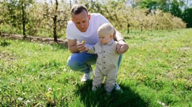Önemseyen baba küçük oğlunun arkasında oturur çimlerin üzerinde durur. Sevgi dolu baba oğluyla konuşur, elindeki çiçeği koklar ve gülümser..