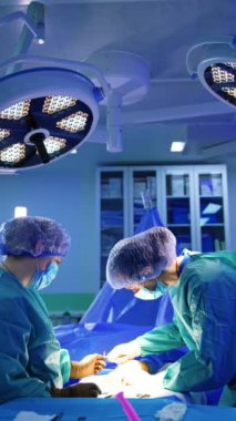 Kadın bir cerrah tarafından yapılan operasyonu parlak yuvarlak lamba aydınlatıyor. Doktorlar ameliyatta metal aletler kullanırlar. Dikey video.