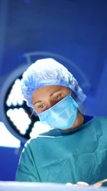Kafkasyalı mavi gözlü doktor ameliyat bölgesine dikkatle bakıyor. Ameliyat yapan kadın cerrahın odaklanmış görüntüsü. Dikey video.