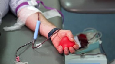 Kırmızı kauçuk kalp, erkek eline sıkışıyor ve kanını bağışlıyor. Kapatın. Arkaplanda kanlı plastik konteynır var..