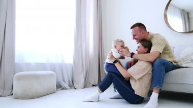 Yerde oturan bir kadın, tatlı bebeğini dizlerinin üstünde kaldırıyor. Baba eşinin arkasında oturup çocuğuna gülümsüyor..