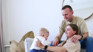 Anne küçük oğluyla oynuyor. Baba arkada oturuyor ve çocuğun elini ağzından çıkarıyor. Aile evde..