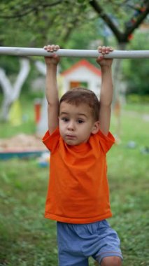 Yatay çubukta sallanan tatlı bir oğlan çocuğu. Çıplak ayaklı çocuk bahçedeki spor envanterinde yuvarlanmaya çalışıyor. Dikey video.
