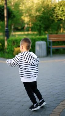 Enerjik çocuk parkta neşeyle zıplıyor. Çocuk scooterına biniyor ve keyifli bir şekilde ara sokakta geziyor. Dikey video.