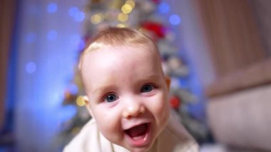 Kamerayı takip eden güzel gülümseyen beyaz gözlü bebek. Kapatın. Çocuk yerde sürünür durur. Noel ağacı ve arka planda siyah bir kedi.