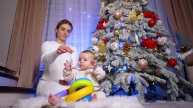 Güzel, beyaz, sarışın kadın bebeğiyle birlikte yerde oturuyor. Annem oğluna Noel ağacından bir kar tanesi süsü veriyor. Düşük açı görünümü.