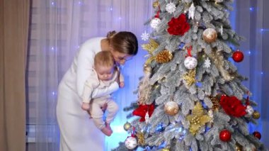 Beyaz elbiseli bir kadın Noel ağacının yanında duran bir bebeği tutuyor. Anne, çocuğun ayağıyla ağaçtaki toplardan birini tekmele..