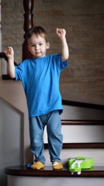 Koyu saçlı, mavi tişörtlü çocuk merdivenlerde duruyor. Çocuk ellerini sallıyor ve kameraya bakarak yere düşüyor. Dikey video.