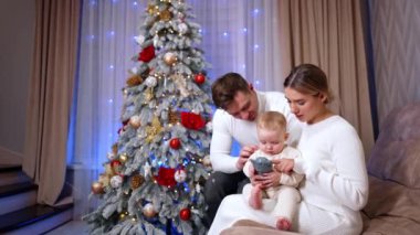 Kanepede oturan anne ve baba sevgili bebeklerine odaklanmış durumdalar. Sevimli çocuk bir oyuncak tutuyor ve onu ısırmaya çalışıyor. Arkaplanda Noel ağacı.