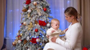Beyaz elbiseli sarışın kadın kanepeye oturup bebek tutuyor. Küçük çocuk ağlamaya başlar ve annesi onu teselli eder. Arkaplanda Noel ağacı.