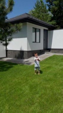 Dışarıdaki yeşil çimlerin üzerinde tişört ve şort giyen üç yaşında bir çocuk duruyor. Neşeli çocuk su tabancasıyla oynuyor. Dikey video.