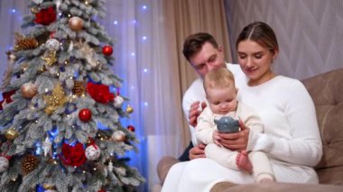 Sevimli küçük bebek elindeki oyuncağa odaklanmış durumda. Ebeveynler sevgili çocuklarını okşar ve öperler. Arkaplanda Noel ağacı.