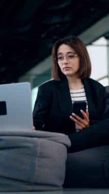 Güzel beyaz kadın, bilgisayarını ve akıllı telefonunu kullanarak fasulye sandalyesinde oturuyor. Düşük açılı bakış açısı işine odaklanmış bir kadın. Dikey video.