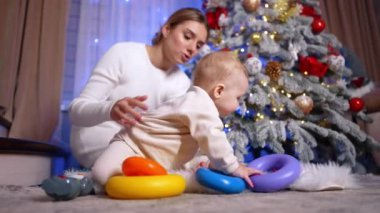 Anne ve küçük bebek Noel ağacının yanında yerde oturuyorlar. Tatlı çocuk piramit halkalarıyla oynuyor. Kara kedi arka planda beliriyor. Düşük açı görünümü.