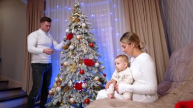 Güzel beyaz bir aile Noel 'i evde kutluyor. Bebekli kadın koltukta otururken, erkek Noel ağacını süslüyor..