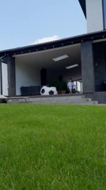 Büyük evin önünde güzel yeşil çimenler. Küçük çocuk kulübenin verandasında oynuyor. Dikey video.