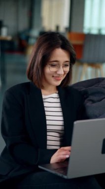 Diz üstü bilgisayar tutan beyaz tenli çekici bir esmer. Gülümseyen kadın bilgisayarında rahat koşullarda çalışıyor. Dikey video.