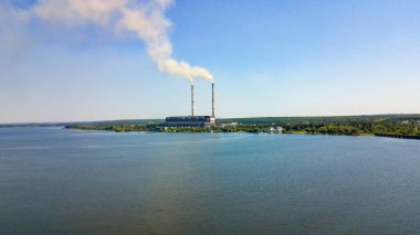 Sakin bir gölün kenarındaki endüstriyel bacalardan duman yükseliyor. Endüstriyel bacalar berrak bir gökyüzünün altında sakin bir göl kenarında duman yayar..