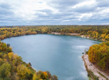 Gün ışığında canlı ağaçlar tarafından çerçevelenmiş sakin bir sonbahar gölü. Sakin bir göl bulutlu bir gökyüzünün altında renkli sonbahar yapraklarını yansıtır.