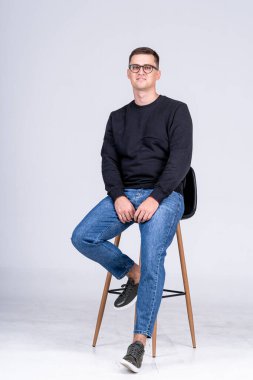 Gözlüklü genç adam parlak bir stüdyoda bir taburede oturuyor. Gözlüklü genç bir adam kendinden emin bir şekilde taburede poz verir.