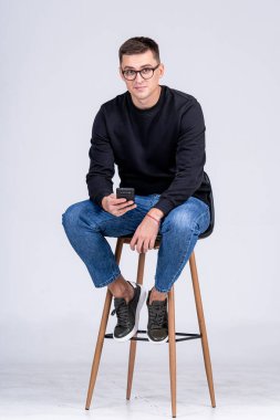 Genç adam stüdyo ortamında akıllı telefon kullanarak tabureye oturdu. Genç bir adam tesadüfen bir taburede oturur.