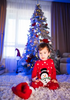 Şenlikli pijamalı çocuk süslü Noel ağacının yanındaki halıda oturuyor. Şenlikli pijama giymiş genç bir çocuk rahat atmosferin tadını çıkarıyor.