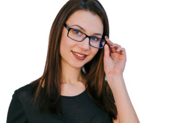 Gözlüklü genç kadın içeride göz giysisini ayarlarken gülümsüyor. Uzun siyah saçlı genç bir kadın, gözlüğünü düzeltirken kendine güvenerek gülümsüyor. Kapalı mekanda dostça bir tavır sergiliyor..