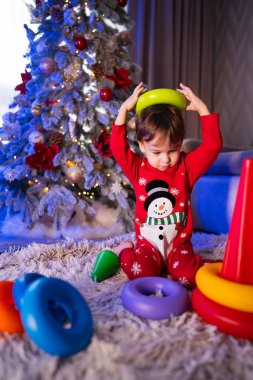 Süslü bir Noel ağacının yanında renkli oyuncaklarla oynayan bir çocuk. Bayram süslemeleriyle çevrili renkli yüzükleri istiflemekten zevk alan neşeli pijamalı bir çocuk..