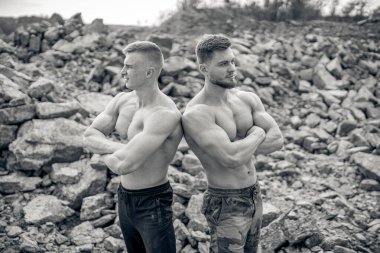 İki kaslı adam kayalık bir açık havada sırt sırta poz veriyor. Kaslı iki adam kayalık arazide güvenle sırt sırta dururlar.