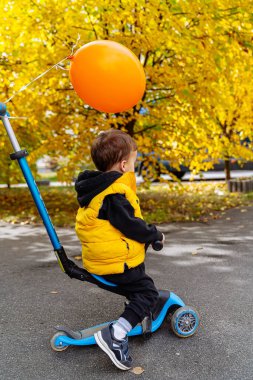 Bir çocuk sonbaharda turuncu bir balonla motosiklet sürmekten hoşlanır. Küçük bir çocuk elinde turuncu bir balonla scooter kullanıyor. Etrafı canlı sonbahar yapraklarıyla çevrili..
