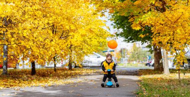 Güneşli bir sonbahar öğleden sonrasında elinde balonla scooter süren bir çocuk. Genç bir çocuk, canlı sonbahar ağaçlarıyla dolu bir patika boyunca scooter sürmekten zevk alır..