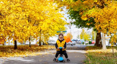 Çocuk, sarı sonbahar yaprakları arasında parkta scooter kullanıyor. Genç bir çocuk, canlı düşen yapraklarla çevrili bir scooterla parkı keşfediyor..